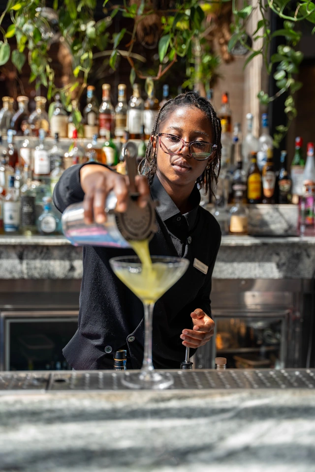 Au bar, une étudiante verse un cocktail dans le verre, bouteilles alignées derrière elle.