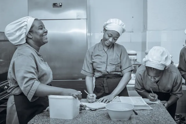 Trois étudiantes en toque prennent des notes en cuisine, l’une éclate de rire